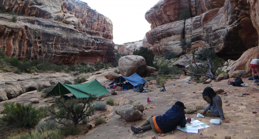Between high canyon walls, people rest at a campsite with two tarp shelters. 